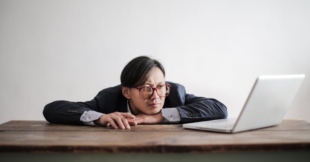 Asian businessman in glasses, resting head on desk, looking at laptop in office setting.